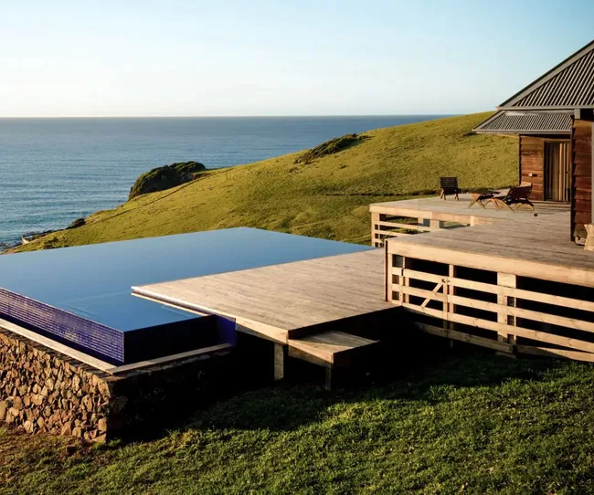 Clifftop house with wooden deck and infinity pool overlooking ocean and grassy hillside under clear blue sky.