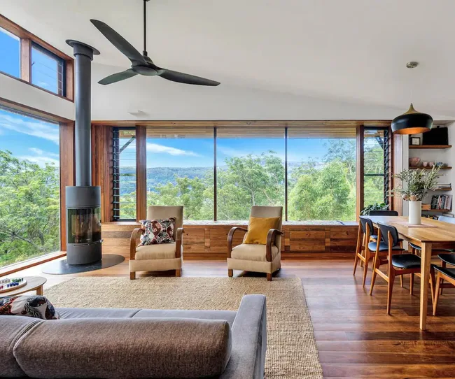 Living room with large windows, forest view, modern furniture, wood floor, ceiling fan, and a cylindrical fireplace.