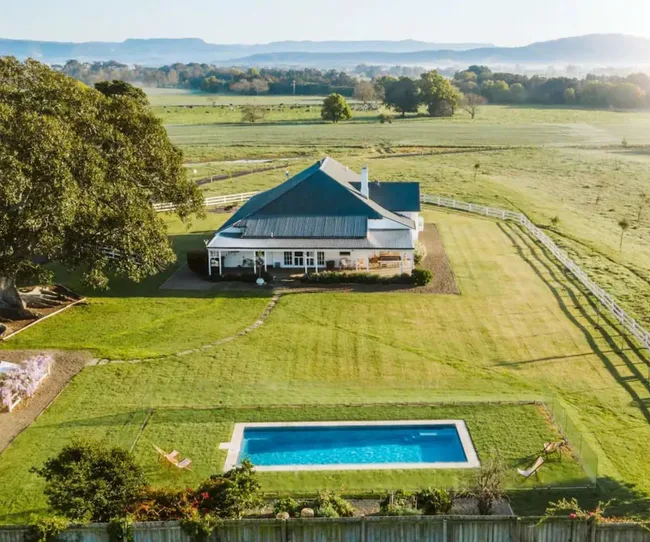 Aerial view of a country house with a blue roof, surrounded by fields, a large tree, and a swimming pool in the backyard.
