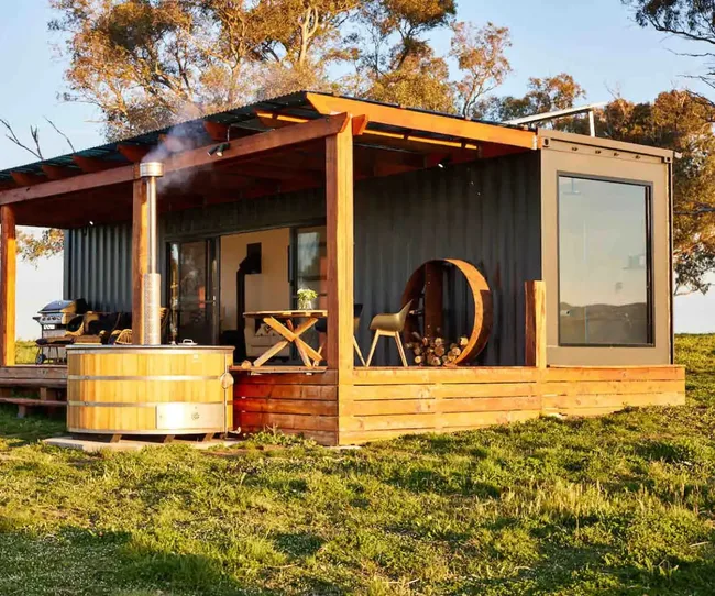 Shipping container cabin with a wooden deck, hot tub, and outdoor seating on grass under a clear sunny sky.