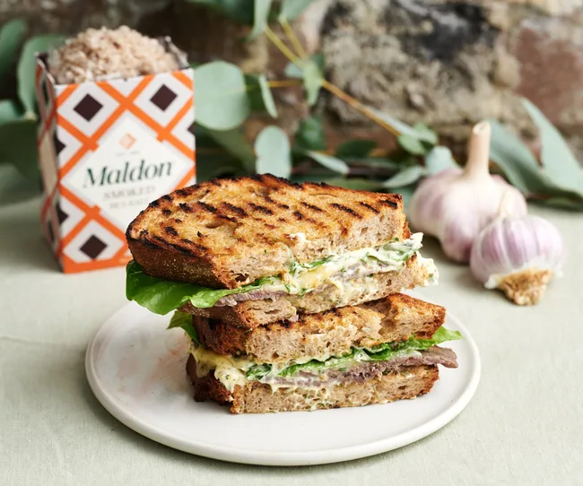 Grilled sandwich with lettuce and spread on a plate, accompanied by Maldon salt and garlic cloves in the background.