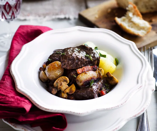 A serving of beef bourguignon with mushrooms, potatoes, and garnishes on a white plate, red napkin beside it.