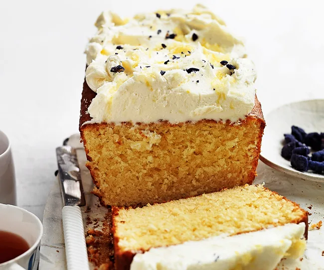 Lemon sponge cake with cream frosting, partially sliced, on a plate with blueberries and a tea cup nearby.