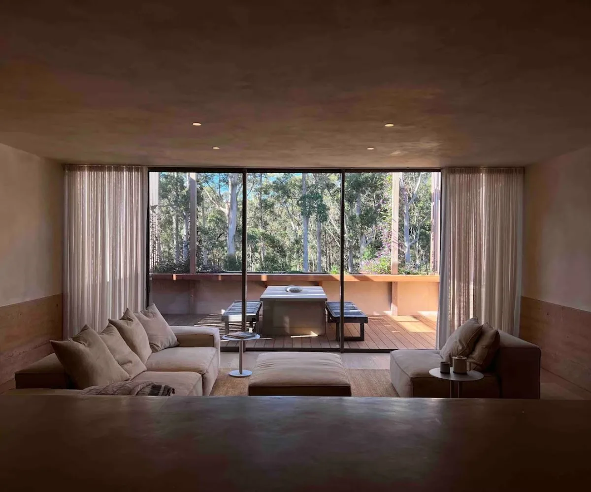 Sunlit living room with beige sectional sofa, large windows, and view of trees outside.