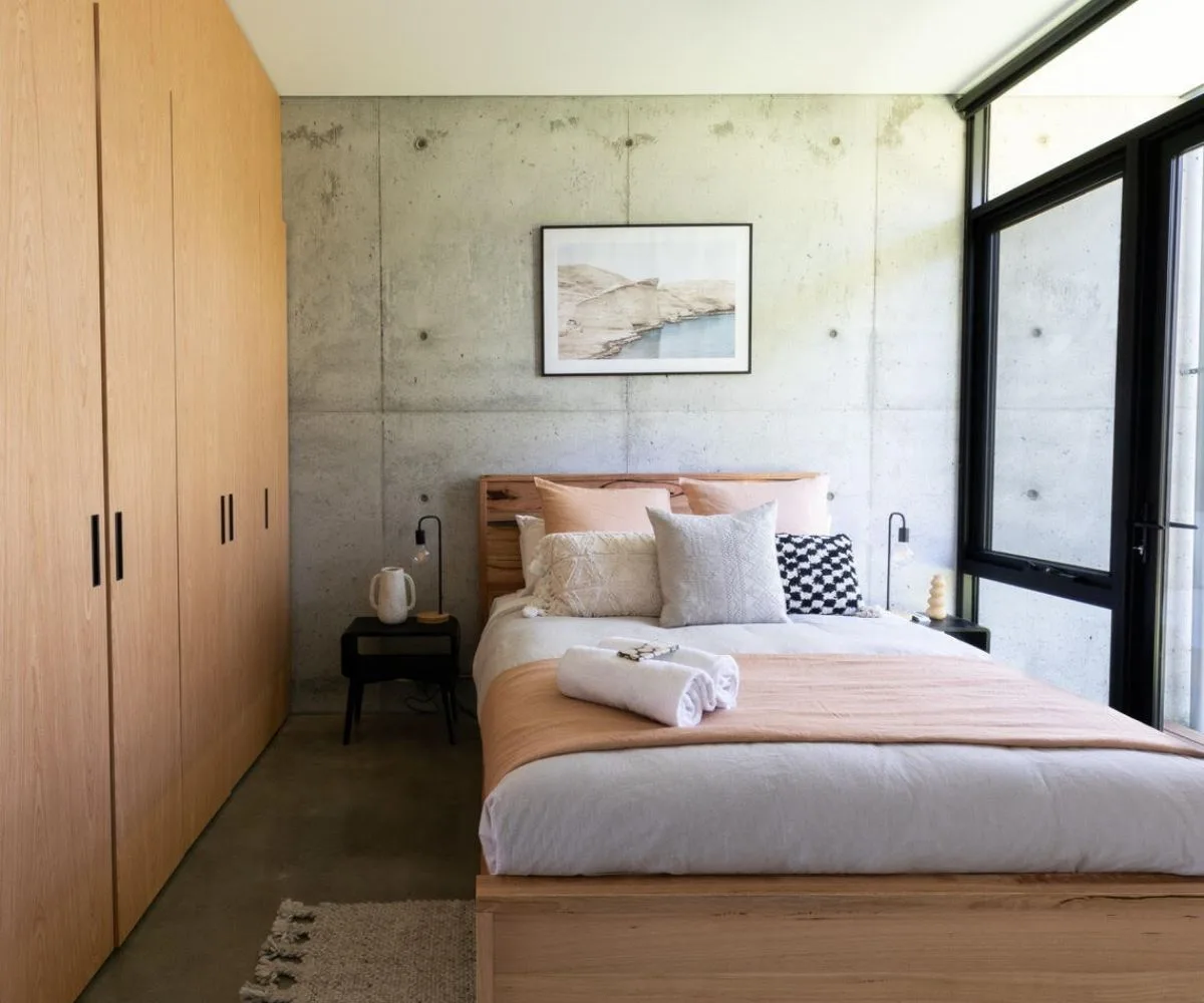 Modern bedroom with concrete walls, wooden wardrobe, bed with beige linens, cushions, and a window with a view.