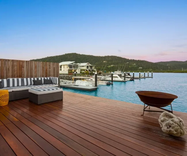 Outdoor deck with striped seating, wooden flooring, fire pit, overlooking marina with boats, under a clear sky.