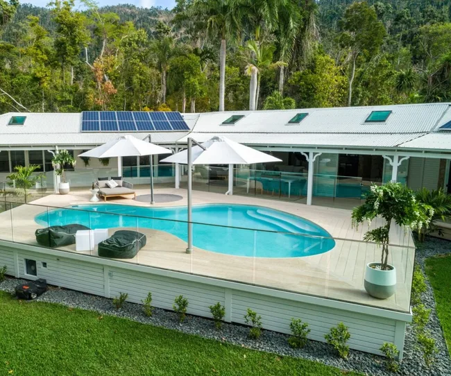 A modern house with a glass-fenced pool, solar panels, and lush green trees surrounding it.