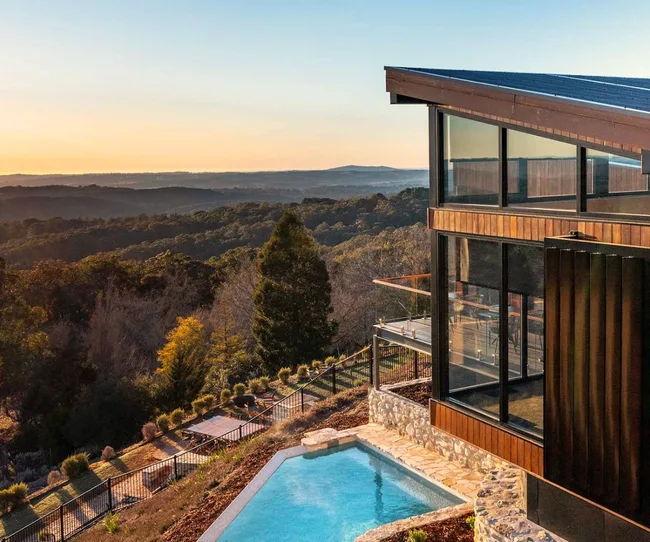 Modern house with large windows overlooking a lush valley at sunset, featuring a pool and a fenced garden.