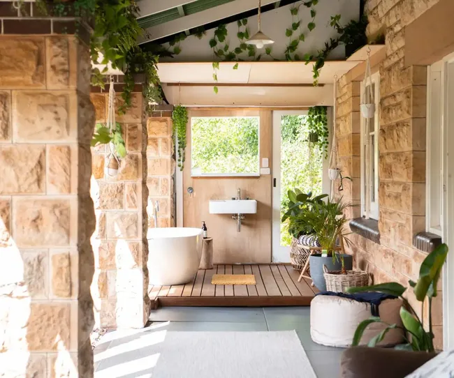 Outdoor bathroom with stone walls, freestanding tub, sink, and lush plants enhancing a natural, serene atmosphere.