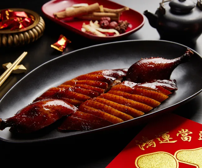 Sliced roast duck on a black plate with traditional Chinese decor and red envelopes on a dark table.