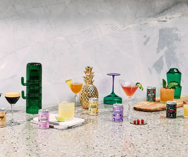 A variety of cocktails in colorful glasses and cans on a marble surface, with decorative elements like a green cactus jug.