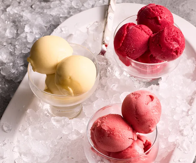 Three glass bowls of sorbet, yellow, pink, and red, placed on a bed of ice.