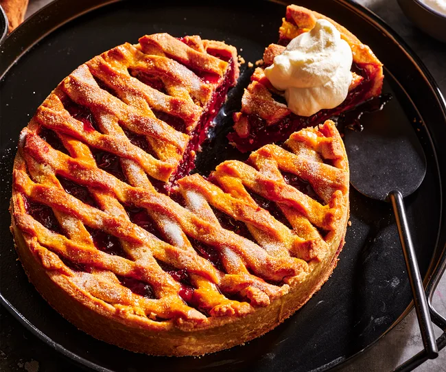 Cherry pie with lattice crust on a black plate, one slice removed and topped with whipped cream.