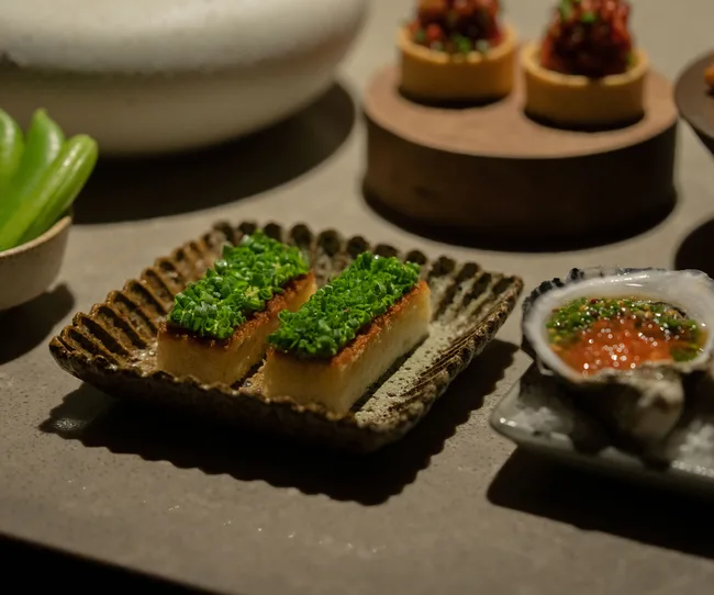 Elegant plate of appetizers with rectangular pieces topped with chopped greens, surrounded by other small dishes.