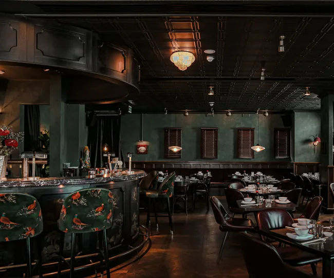 Elegant restaurant interior with a curved bar, floral chairs, dark green walls, and tables set for dining.