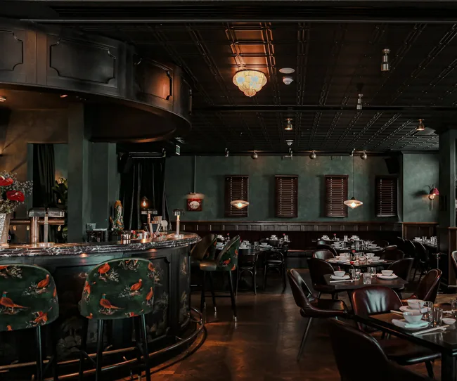Elegant restaurant interior with a curved bar, floral chairs, dark green walls, and tables set for dining.