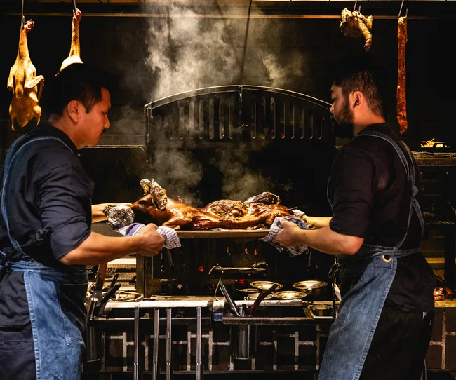 Two chefs in aprons roast a whole pig in a smoky kitchen, with hanging poultry in the background.