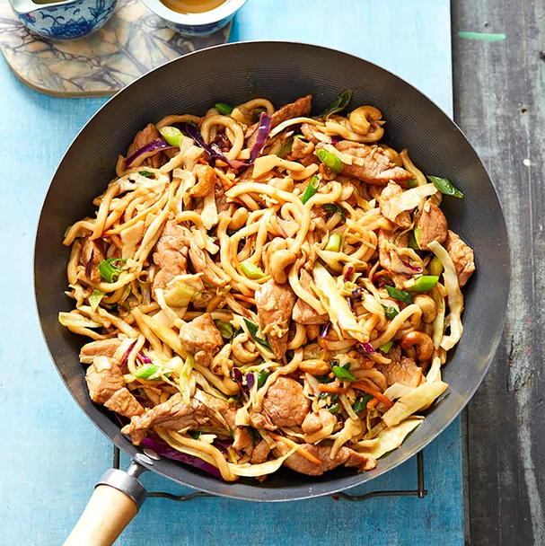 Wok filled with pork stir-fry, noodles, and vegetables on a blue surface.
