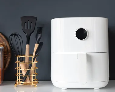 A white air fryer next to kitchen utensils, a cutting board, and a jar of lentils on a countertop.
