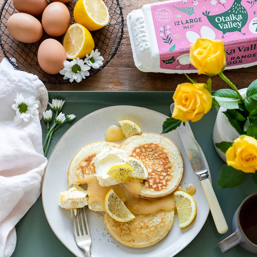 Plate of pancakes with lemon slices and cream, surrounded by eggs, lemons, and yellow roses on a wooden table.