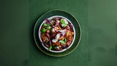 A bowl of roasted vegetables and greens drizzled with a white sauce on a green textured tablecloth.