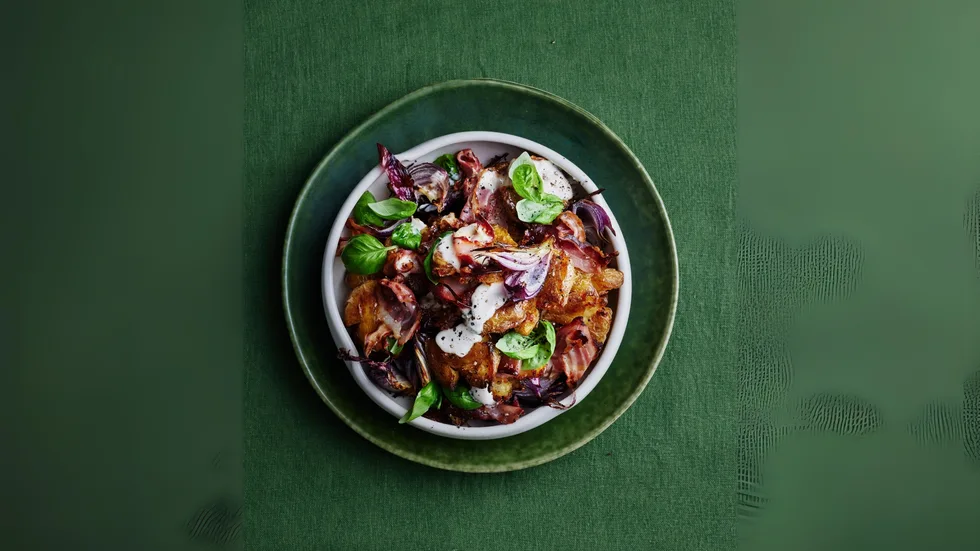A bowl of roasted vegetables and greens drizzled with a white sauce on a green textured tablecloth.
