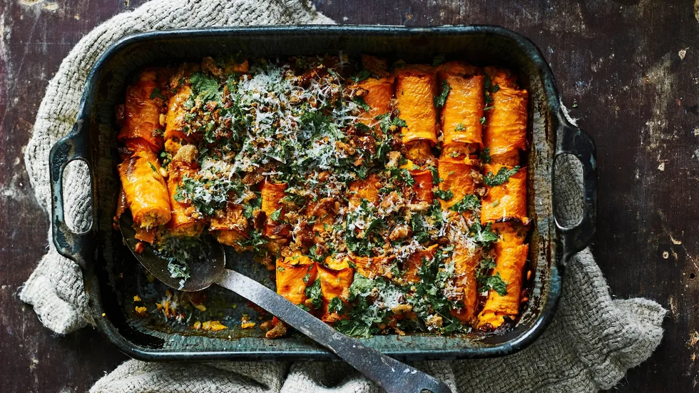 A rustic dish of baked pasta rolls with cheese and herbs, garnished with breadcrumbs in a dark baking dish on a cloth.