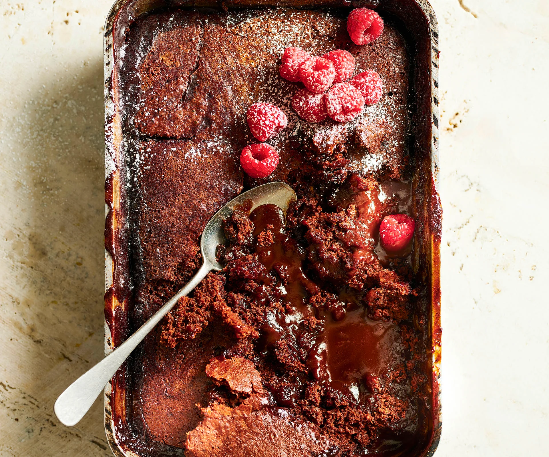 Hot chocolate pudding in a baking dish with a spoon, topped with fresh raspberries and powdered sugar.