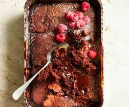 Hot chocolate pudding in a baking dish with a spoon, topped with fresh raspberries and powdered sugar.