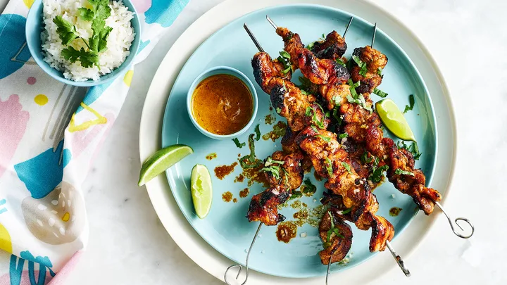 Grilled chicken skewers on a plate with dipping sauce, lime wedges, and a bowl of rice garnished with cilantro.