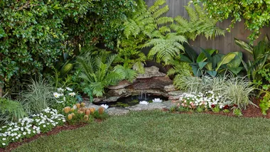 Small backyard pond in a corner of the garden surrounded by flowering plants.