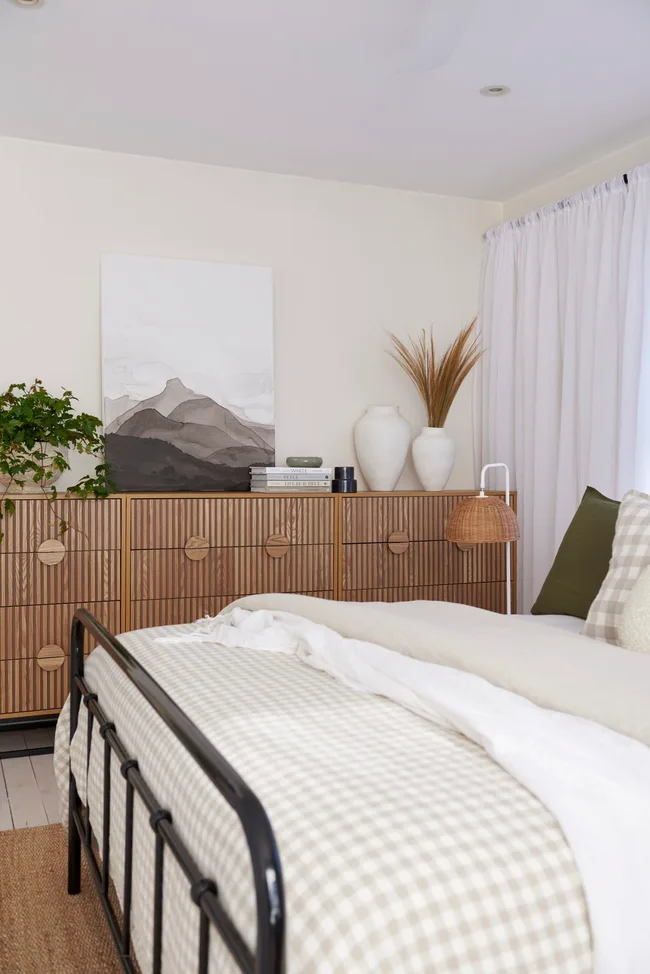 side on shot of bedroom with metal black bed frame and beige quilt cover folded over on the bed. sideboard in the background with a lamp and various trinkets on it