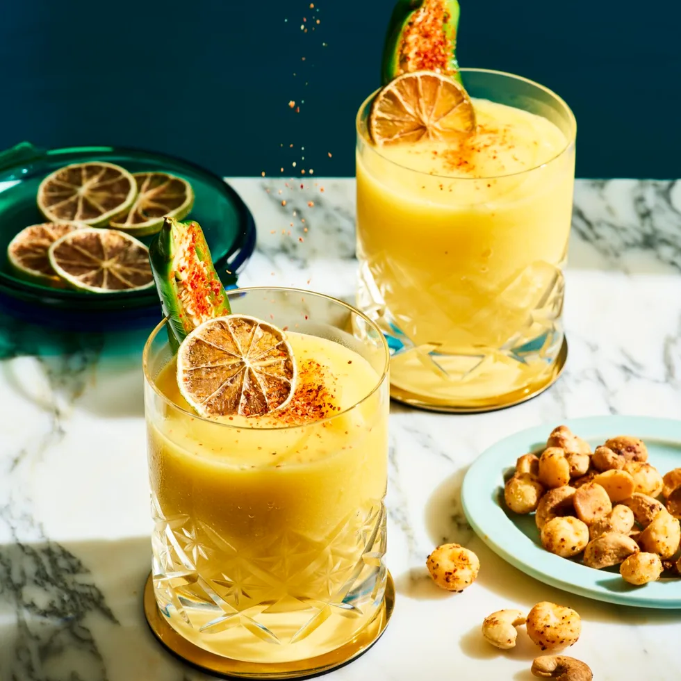 2 short tumbler style glasses with a mango liquid inside, garnished with a green jalapeno and dried lime. A ladies hand is hovering above squeezing lime into the cocktails. The cocktails are on a marble table top and navy blue background.