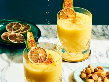 2 short tumbler style glasses with a mango liquid inside, garnished with a green jalapeno and dried lime. A ladies hand is hovering above squeezing lime into the cocktails. The cocktails are on a marble table top and navy blue background.