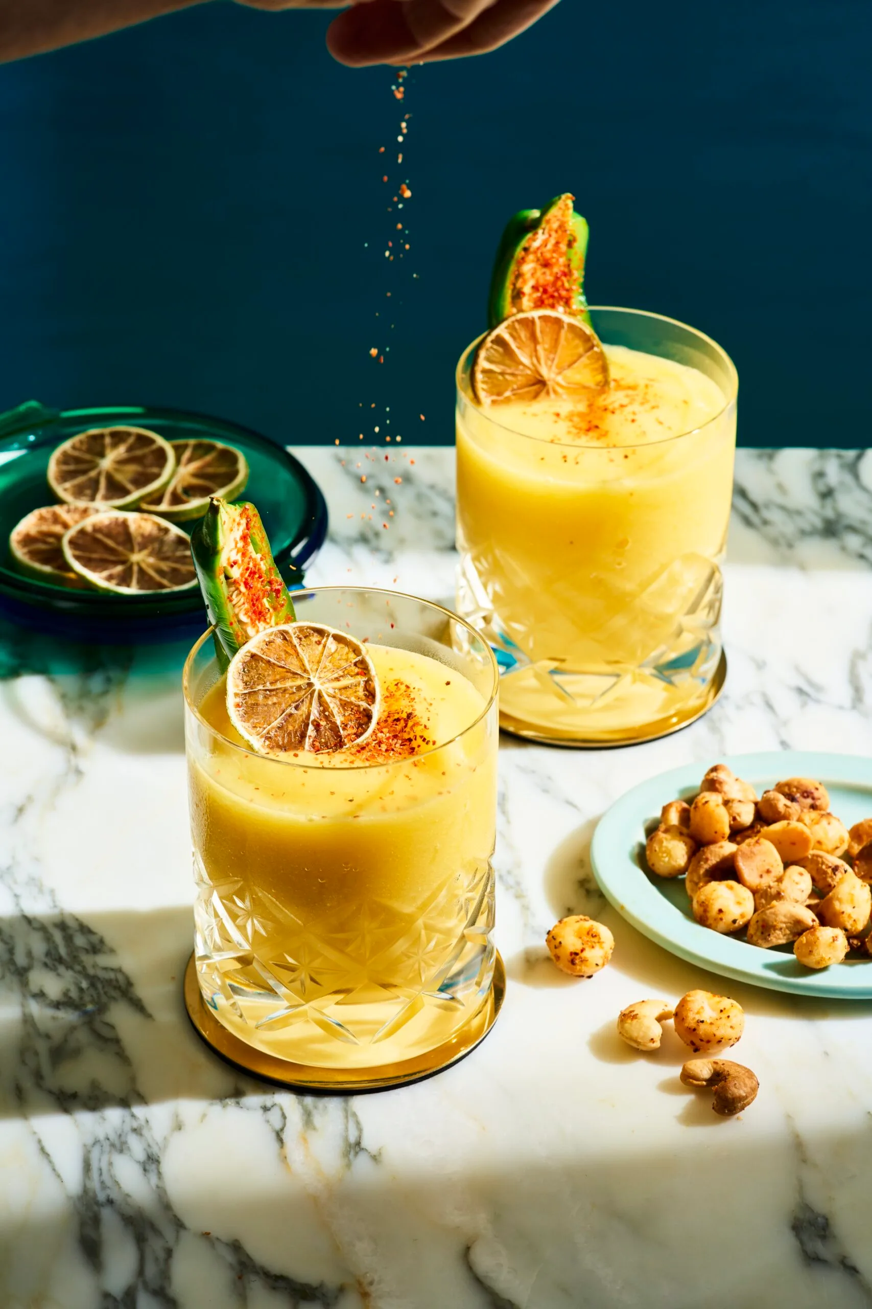 2 short tumbler style glasses with a mango liquid inside, garnished with a green jalapeno and dried lime. A ladies hand is hovering above squeezing lime into the cocktails. The cocktails are on a marble table top and navy blue background.