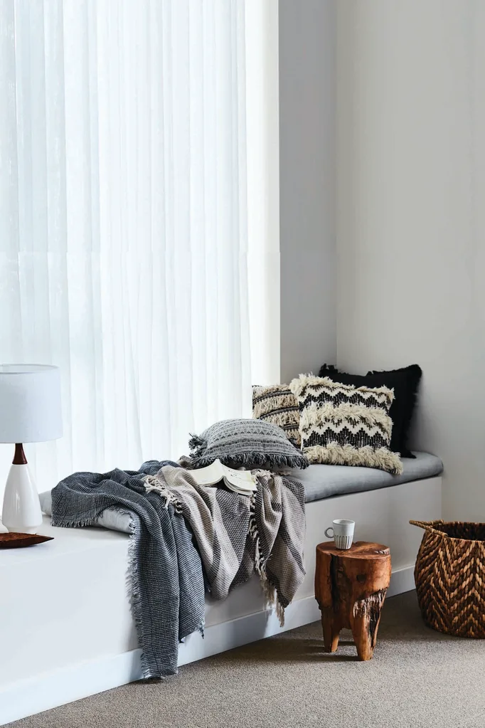 Window seat with sheer curtains, throw blankets and a mug on a timber side table.