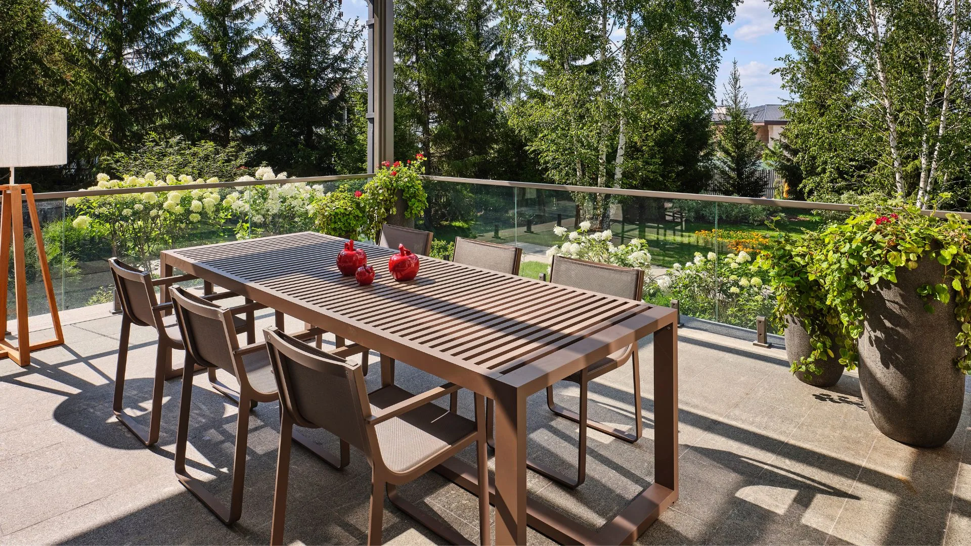 A modern outdoor dining set with a slatted table and chairs sits on a patio under a glass pergola.