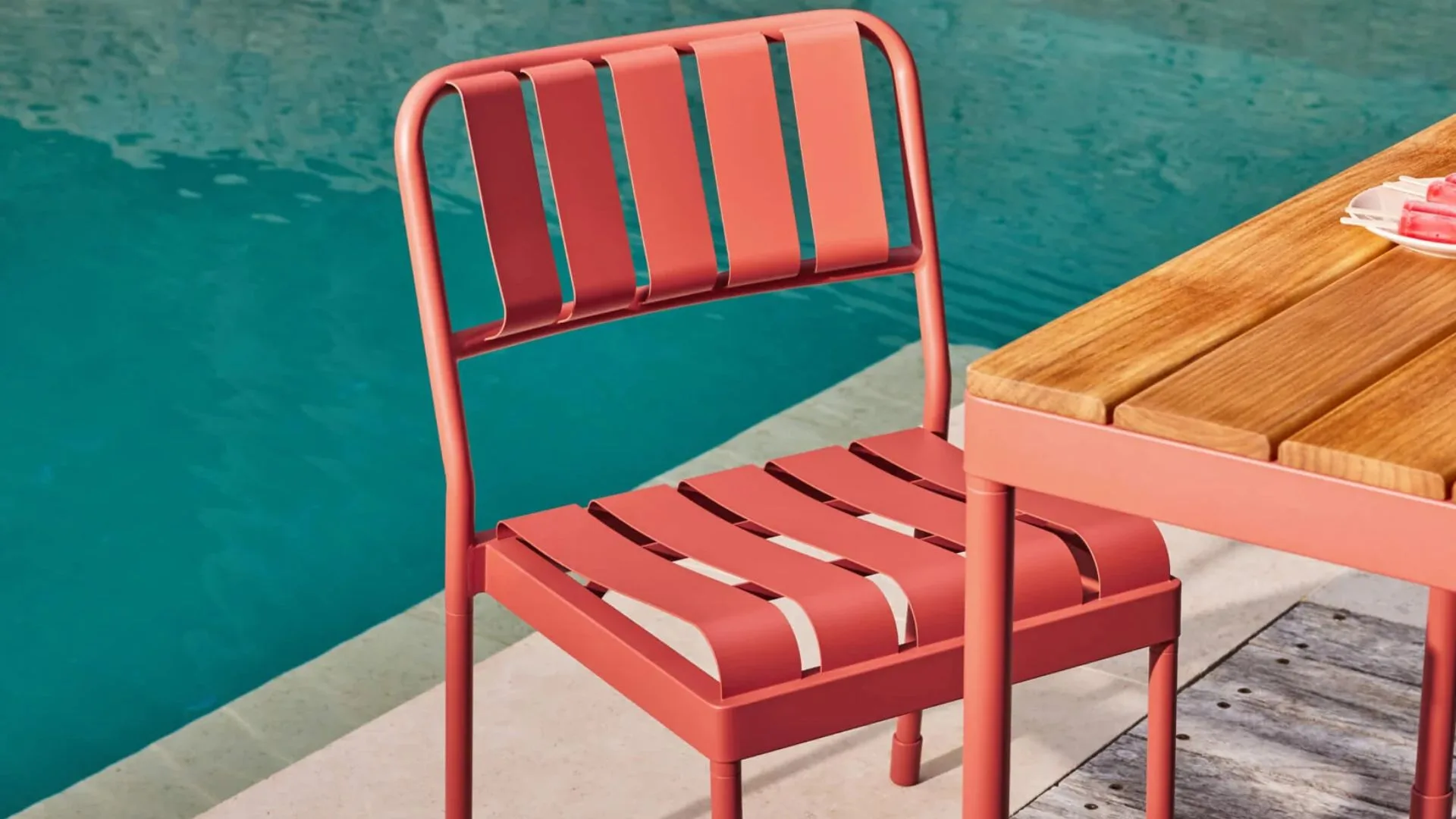 A coral-pink metal chair from the Koala Torakina Outdoor Dining Set sits beside a wooden table by a swimming pool.