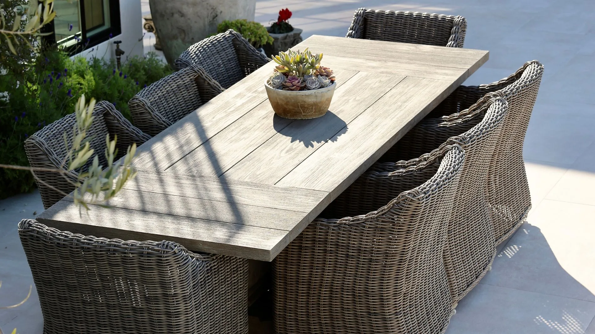 A wooden outdoor dining table is surrounded by woven wicker chairs, with a succulent planter in the centre.