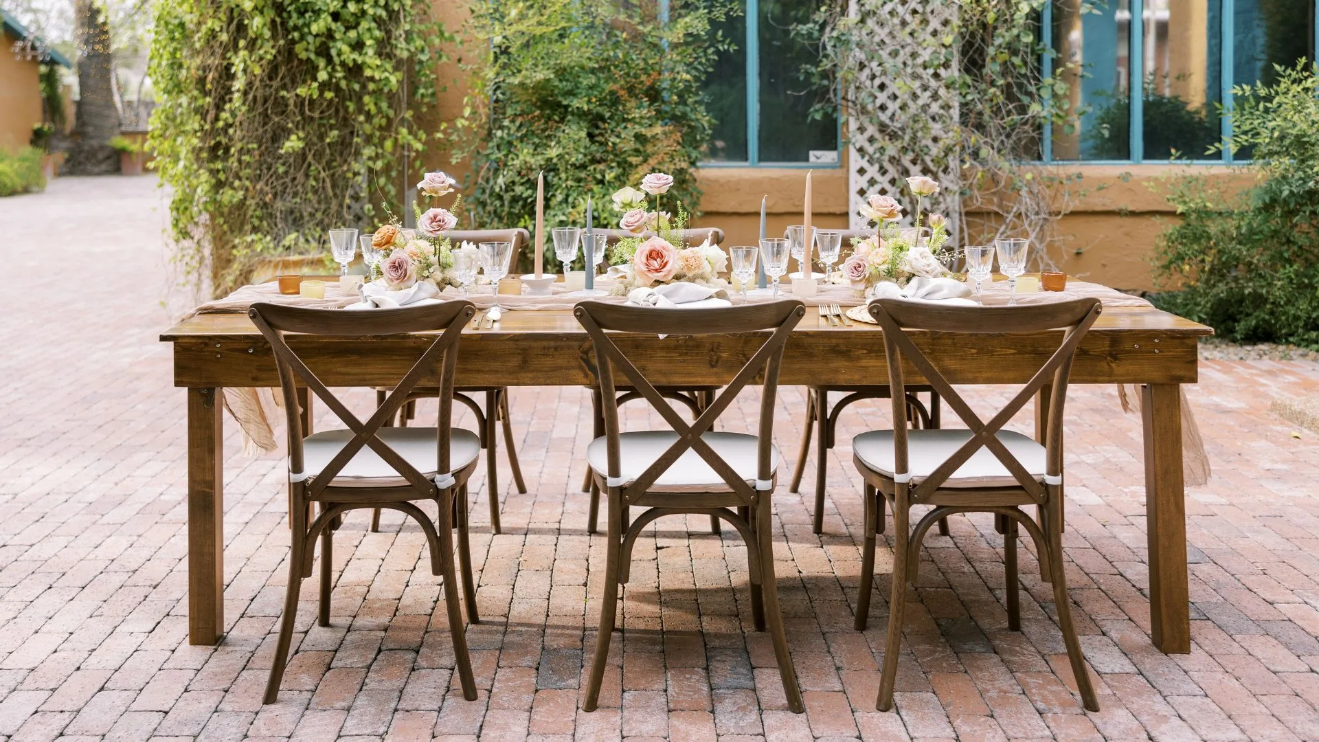A rustic outdoor dining table with cross-back chairs is set on a brick patio with floral arrangements and candles.
