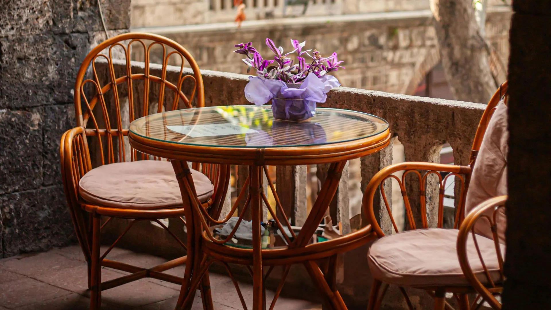 A glass-topped rattan table and two matching chairs with cushions are placed on a stone balcony with a potted plant.
