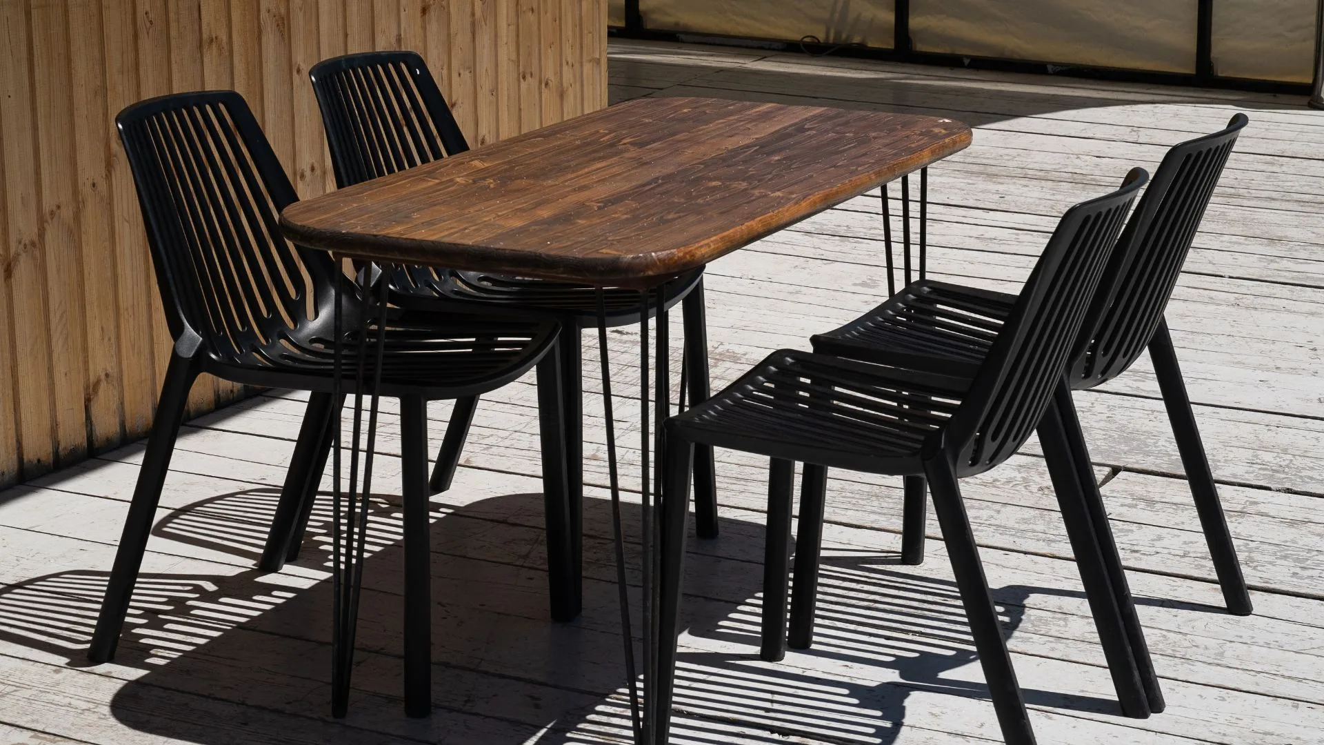 A dark wood table with hairpin legs is surrounded by four black, slatted plastic chairs on a light-coloured wooden deck.