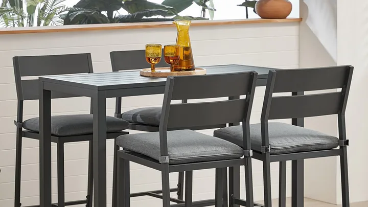Four grey cushioned chairs around a rectangular table with a tray of amber glasses and a jug, set indoors.