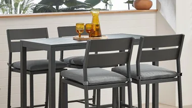 Four grey cushioned chairs around a rectangular table with a tray of amber glasses and a jug, set indoors.