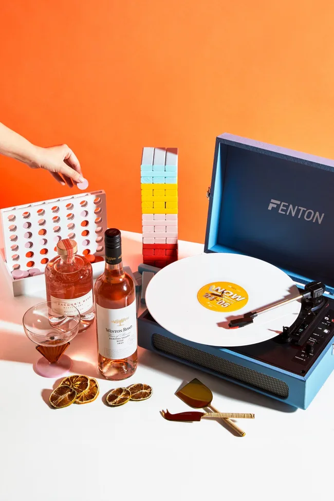 Wine, a record player, board games on a white table against an orange background. A collection of gift ideas for people who love to cook and entertain their friends at home.