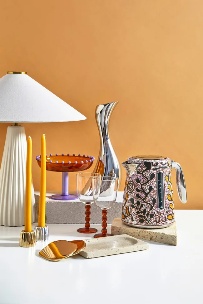 Home decor gifts including a white lamp, taper candles and a Breville kettle arranged on a white table against an orange backdrop.