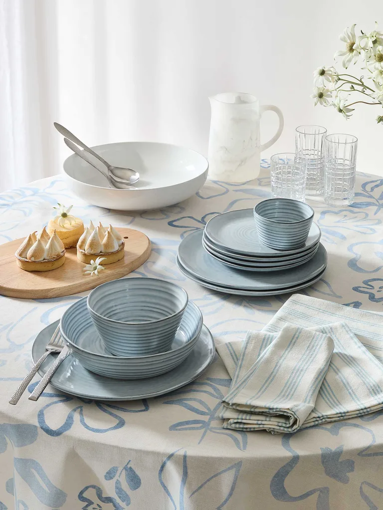 A selection of tableware from Kmart's Feb 2025 homewares collection, featuring a white and blue tablecloth, blue dinner set and white resin serving bowls and jugs.