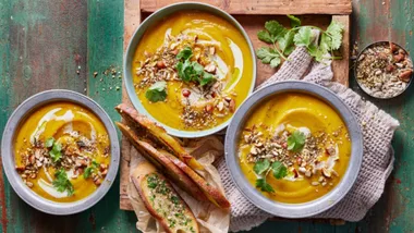 Three bowls filled with pumpkin soup, served with crusty bread and seeds and nuts