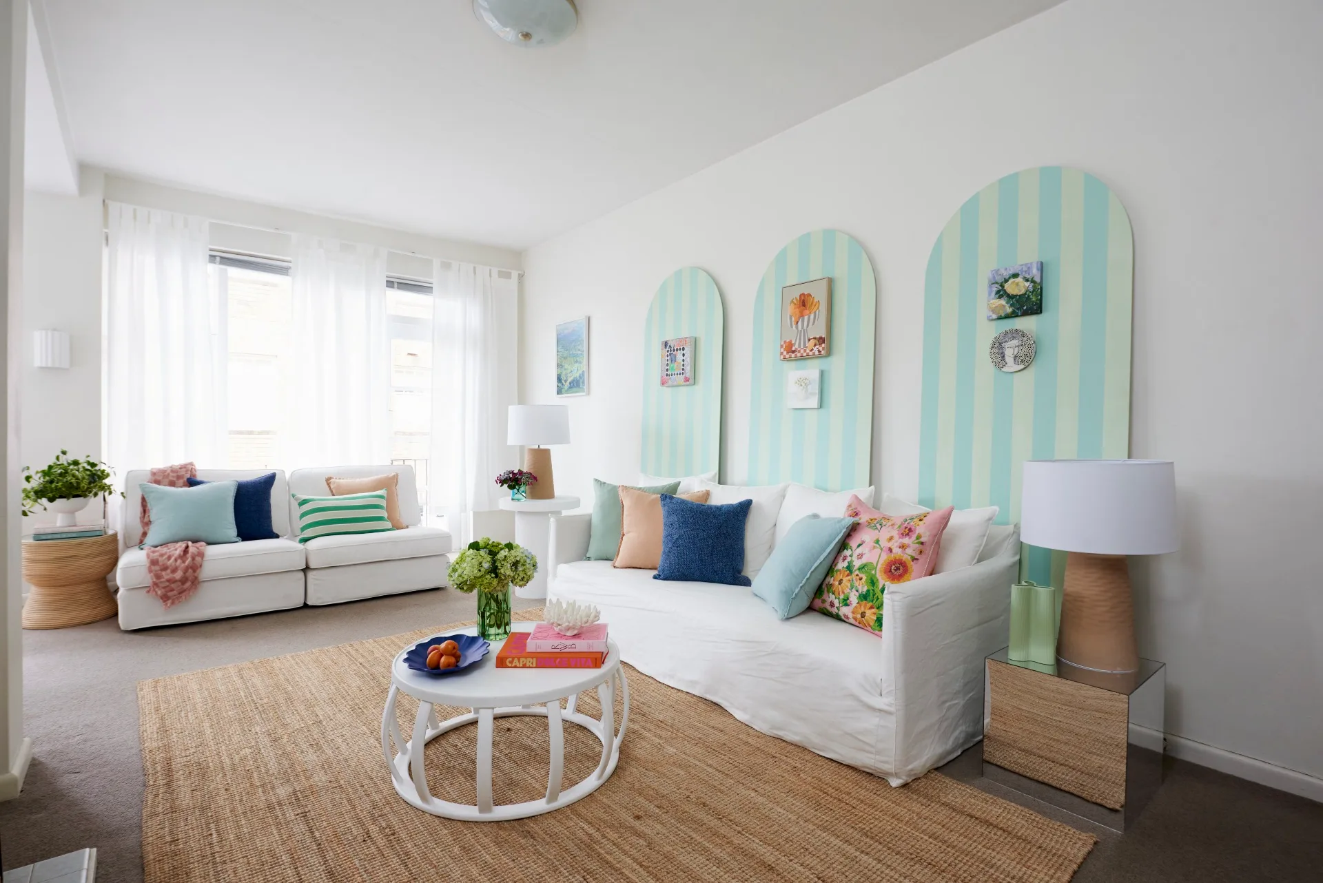 Bright living room with white sofas, colorful cushions, striped decor on walls, coffee table, plants, and lamps.