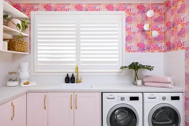 Stylish laundry room with pink cabinets, floral wallpaper, washer and dryer, plants, and decorative items on shelves.